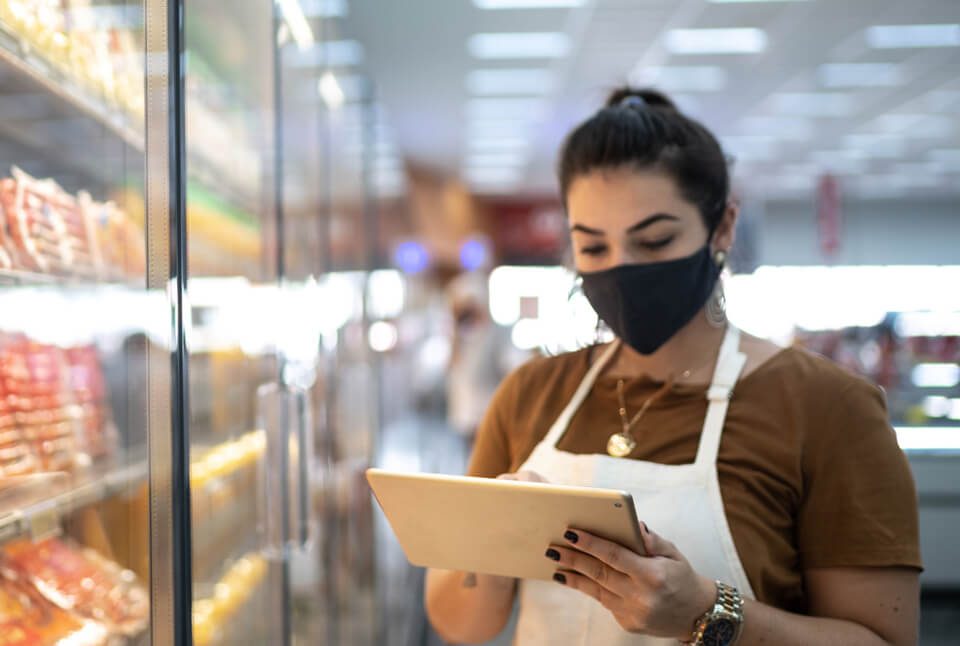 A woman in a mask and apron uses a tablet in a grocery store aisle, standing near refrigerated shelves with packaged goods.