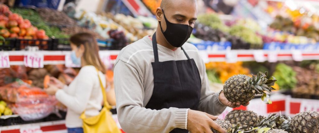 A person wearing a black mask and apron inspects pineapples in a grocery store. A woman in the background looks at vegetables.