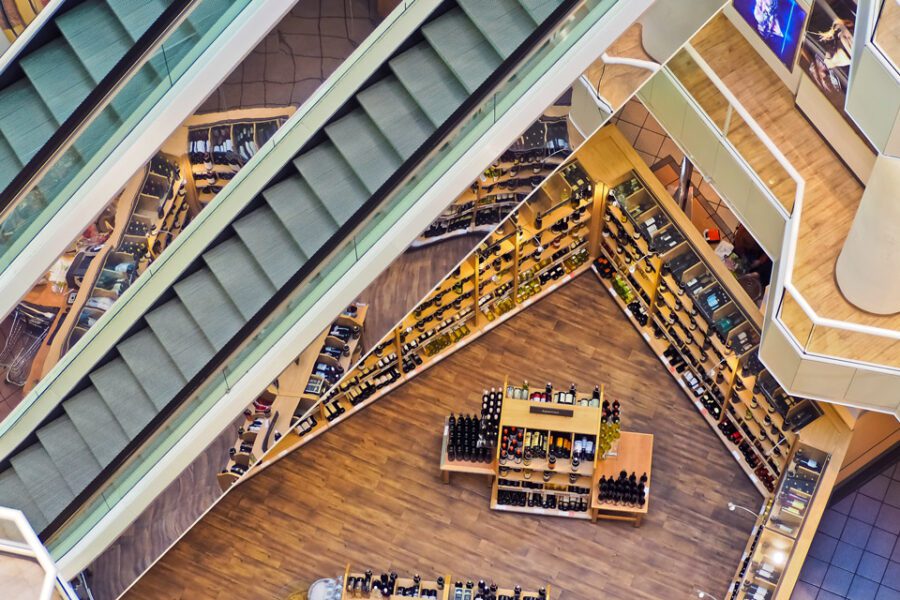 Aerial view of a shopping mall interior with escalators and a shoe store. The shoe store displays various footwear on wooden shelves and a central display table.