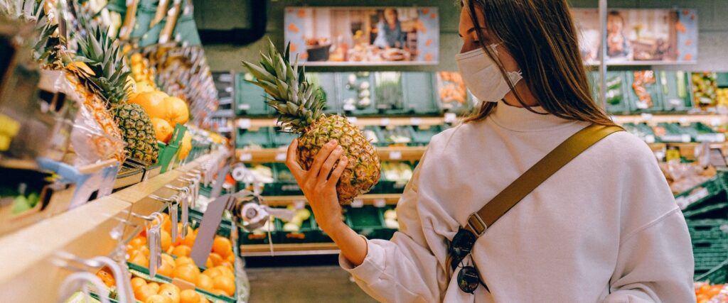Person in a white sweater and face mask holding a pineapple in a grocery store produce aisle.