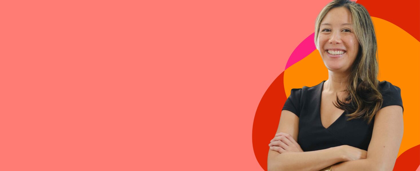 A person with long hair and a black top smiles with arms crossed. The background has abstract shapes in orange, red, and pink tones.