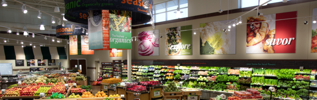A brightly lit grocery store section displaying various fruits and vegetables, with colorful produce banners hanging from the ceiling.