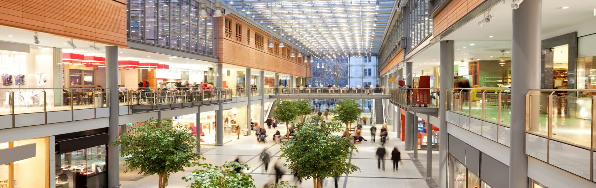 Spacious modern shopping mall with glass ceiling, two-story layout, and various shops. Indoor trees and scattered people create a bustling atmosphere.