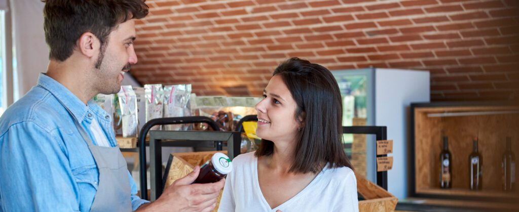 A man in an apron shows a bottle to a smiling woman in a shop with wooden shelves and clay brick ceiling.