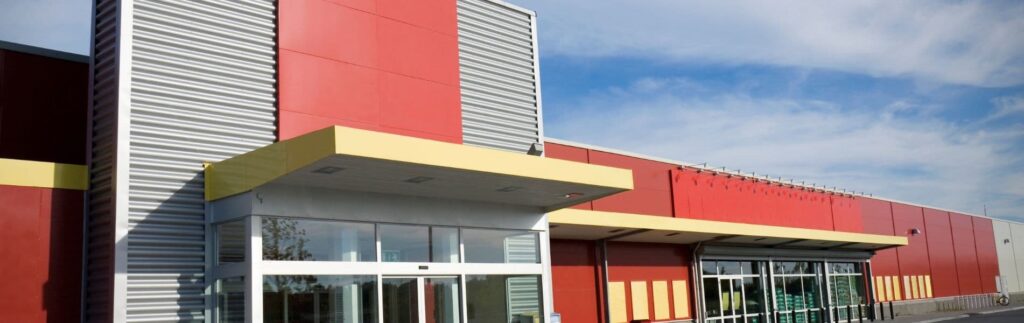 Large red and yellow commercial building with a modern design, featuring a main entrance with glass doors and an adjacent wall with metal paneling under a blue sky.
