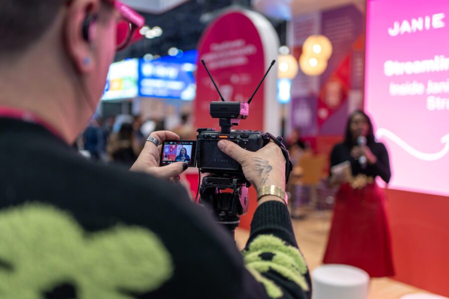 A person holding a camera films a woman speaking on stage at an indoor event with bright lights and colorful displays in the background.
