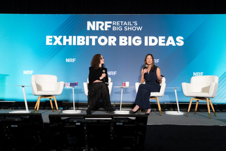 Two women sit on stage in discussion at the NRF Retail’s Big Show, with a blue “Exhibitor Big Ideas” backdrop and empty chairs beside them.