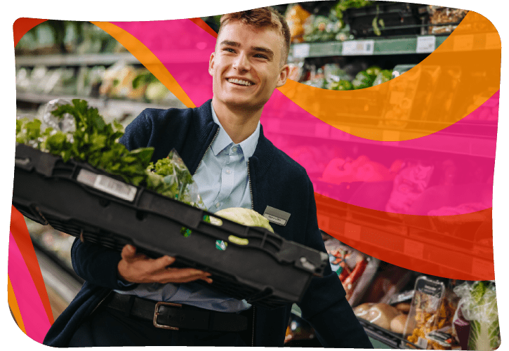 A person in a grocery store holds a crate filled with fresh vegetables, including lettuce and herbs.