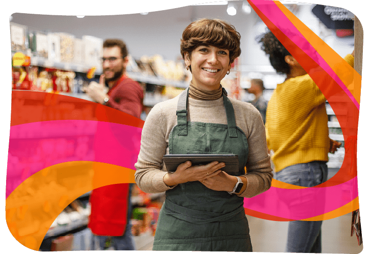 Person in green apron holding tablet in a store aisle, with two people browsing in the background.