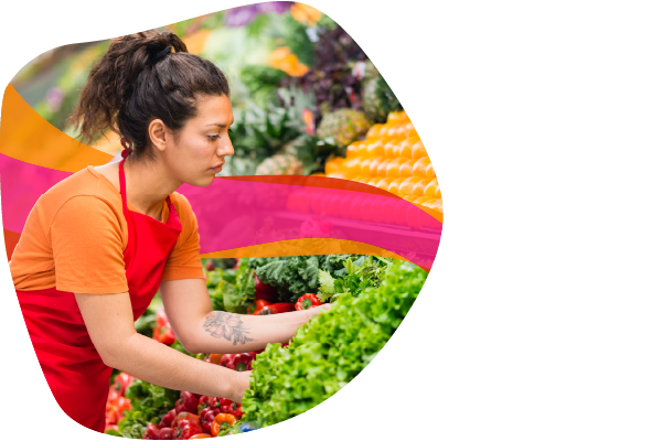A person in an orange shirt and red apron arranging vegetables in a grocery store.