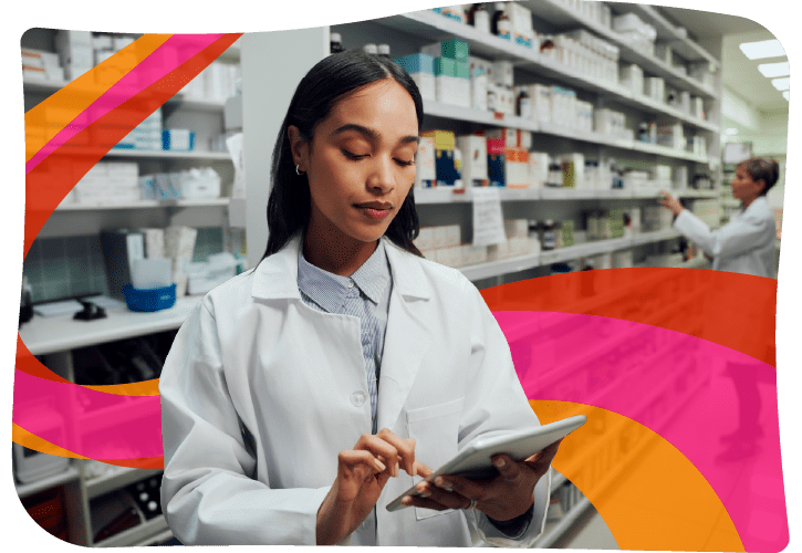 A pharmacist wearing a white coat uses a tablet in a pharmacy, with shelves of medications in the background. Another person is organizing products on the shelves.