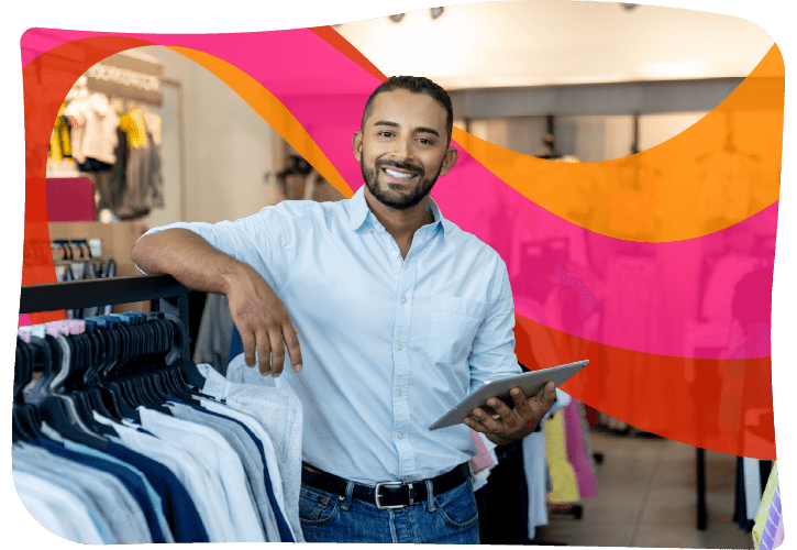 Man in a clothing store, smiling, holding a tablet, standing near a rack of shirts.