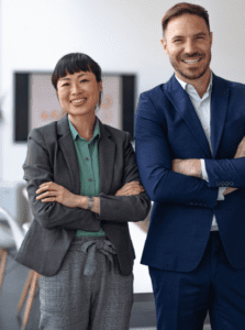 Two smiling people in business attire stand with arms crossed. The woman wears a gray blazer and plaid pants, and the man is in a blue suit. They are indoors with a blurred background.