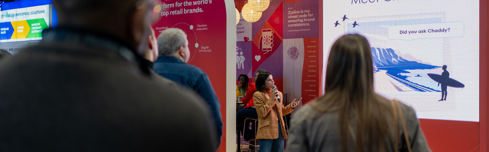 A woman stands with a microphone, gesturing towards a presentation slide. A few people in the audience watch attentively.