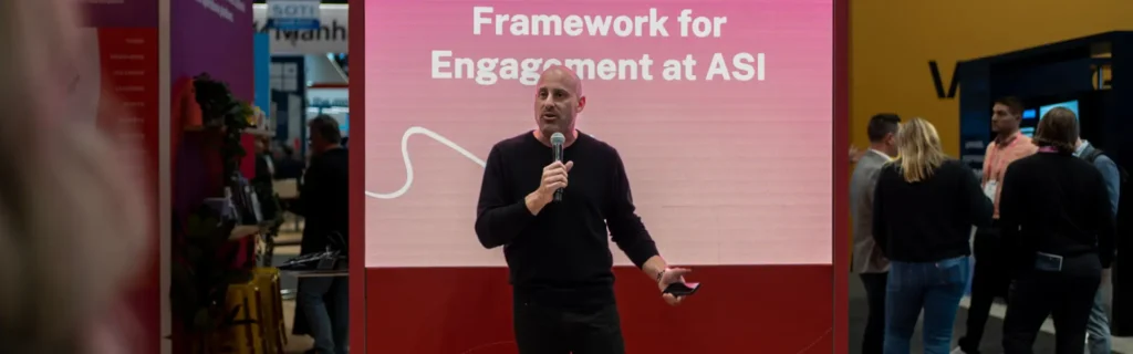 A person speaks into a microphone in front of a screen displaying the text "Framework for Engagement at ASI," with people standing nearby.