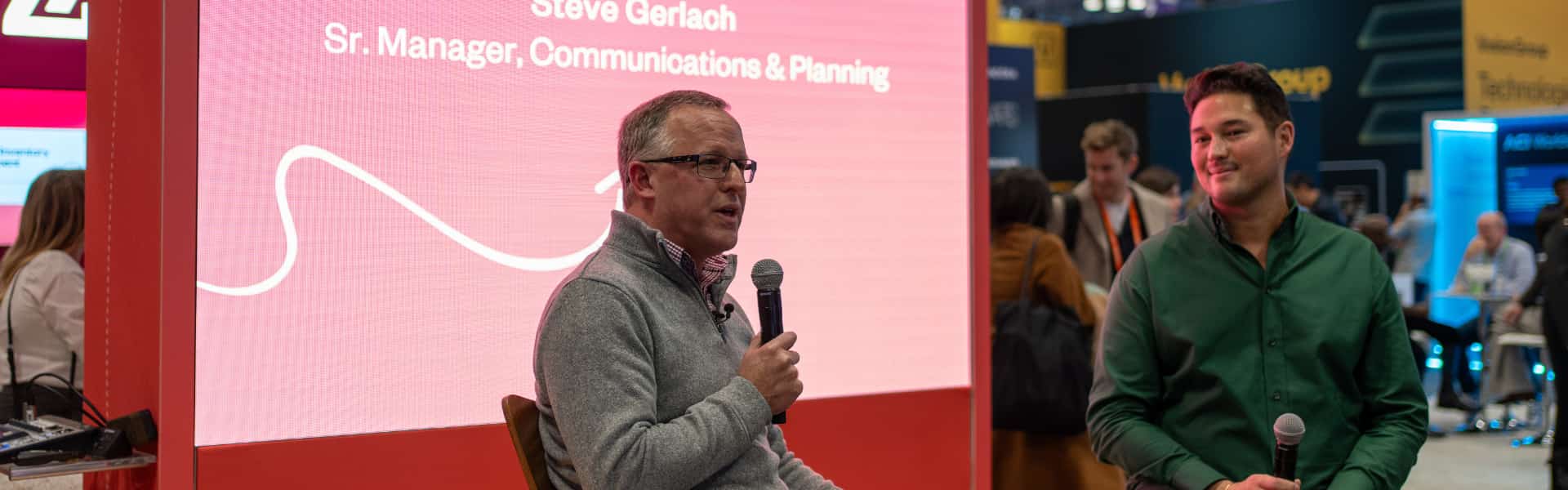 Two men sitting with microphones in front of an audience at a tech conference. The background displays a presentation slide with text about a communications and planning manager.