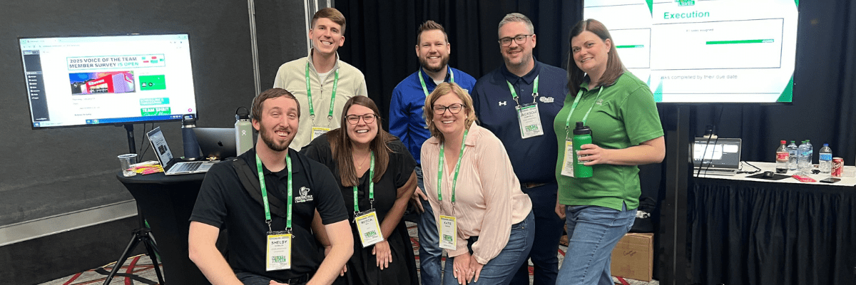 A group of seven people wearing conference badges pose and smile for a photo in front of a booth with monitors and presentation materials.