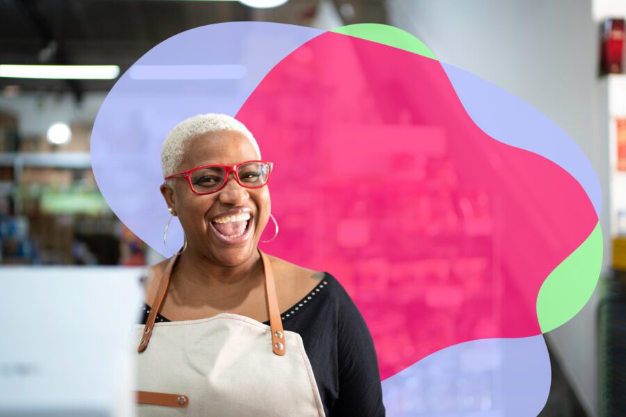 A smiling woman wearing red glasses and an apron stands indoors, with colorful abstract shapes in the background.