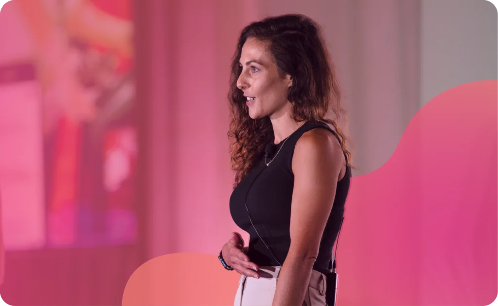 A woman with long curly hair stands on stage, wearing a sleeveless black top and light-colored pants, speaking with a microphone headset against a pink-lit background.