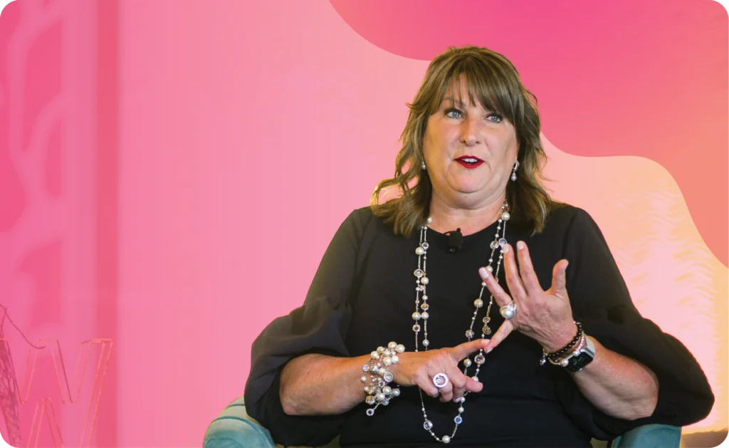 A woman with shoulder-length brown hair and jewelry sits on a chair, gesturing with her hands, against a pink and orange gradient background.