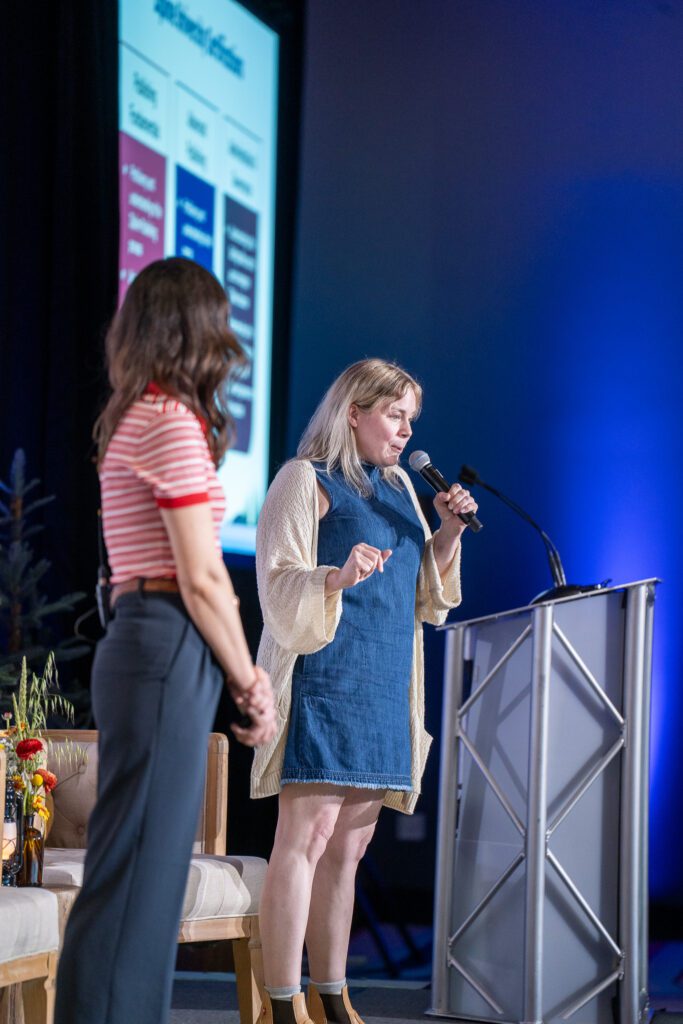 Two women are speaking on stage at a conference; one stands at a podium holding a microphone while the other stands nearby. A presentation slide is displayed in the background.