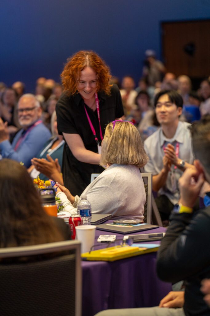 A woman with curly red hair smiles and shakes hands with a seated attendee at a conference, while others watch and applaud.