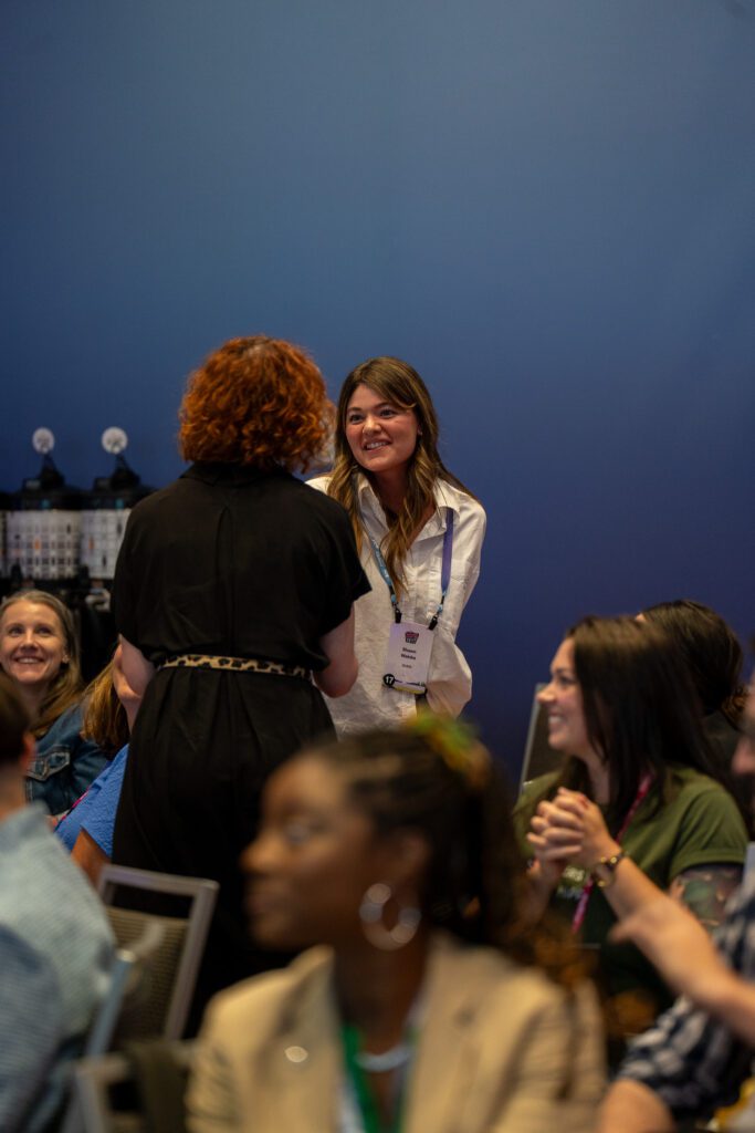A woman in a white shirt stands and smiles while interacting with another woman in a black outfit during an indoor event, with seated attendees in the background.