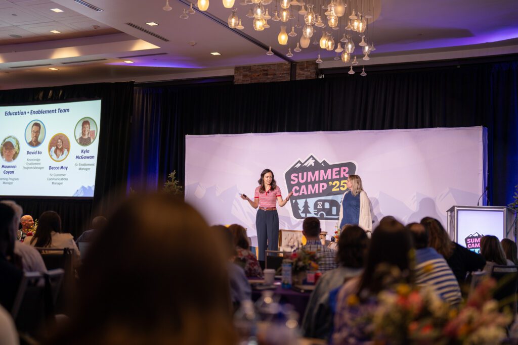 Two women stand on stage giving a presentation at a conference called "Summer Camp '25" while attendees listen from the audience.
