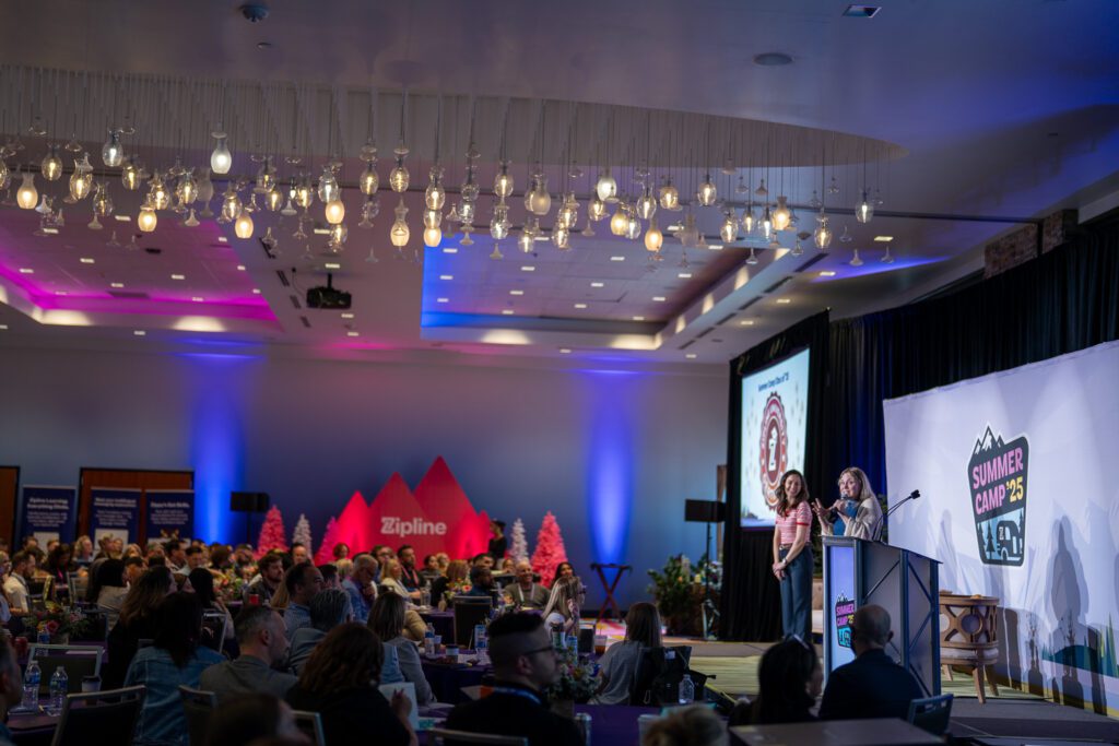 Two speakers stand at a podium addressing an audience in a conference room with colorful lighting and a "Summer Camp '23" banner on stage.