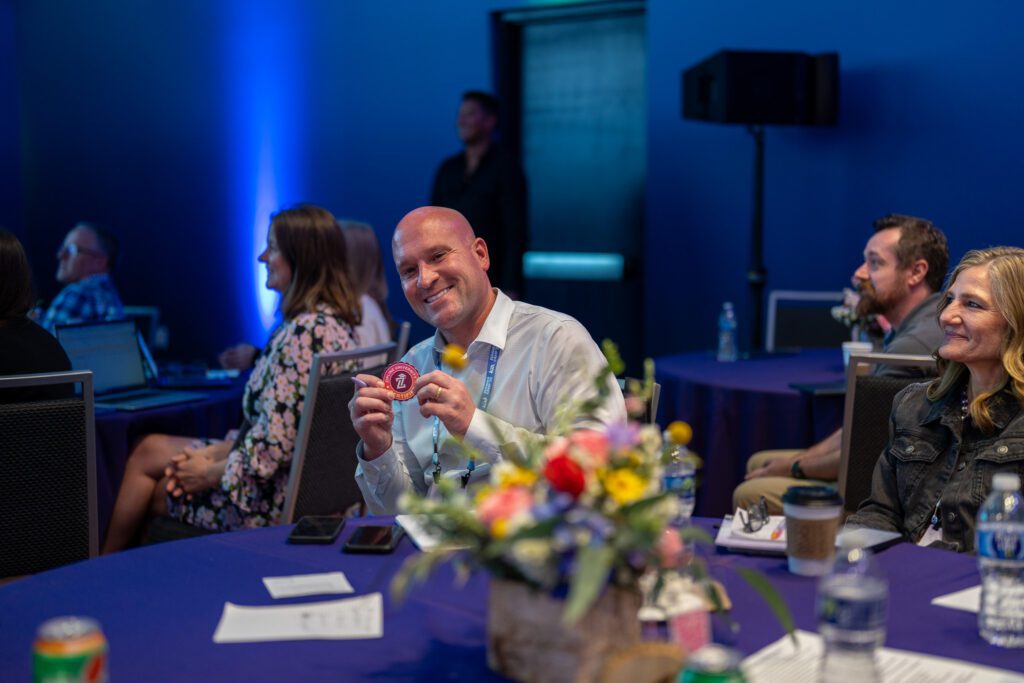 A man seated at a table in a conference room holds up a round object and smiles at the camera. Other people are seated nearby, and there are flowers and drinks on the table.