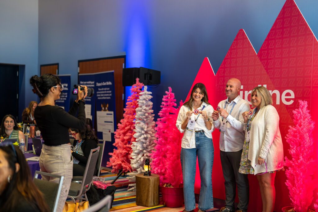 Three people pose for a photo in front of pink trees and a red backdrop at an indoor event while another person takes their picture.