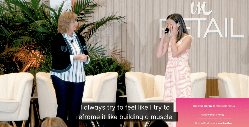 Two women stand on a stage with white chairs and plants. One woman speaks while the other covers her face. Text overlay reads, "I always try to feel like I try to reframe it like building a muscle.