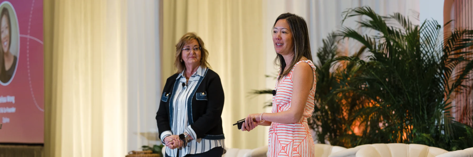 Two women stand on a stage; one is speaking while the other looks on. A screen with a photo and text is partly visible in the background, along with plants and draped curtains.