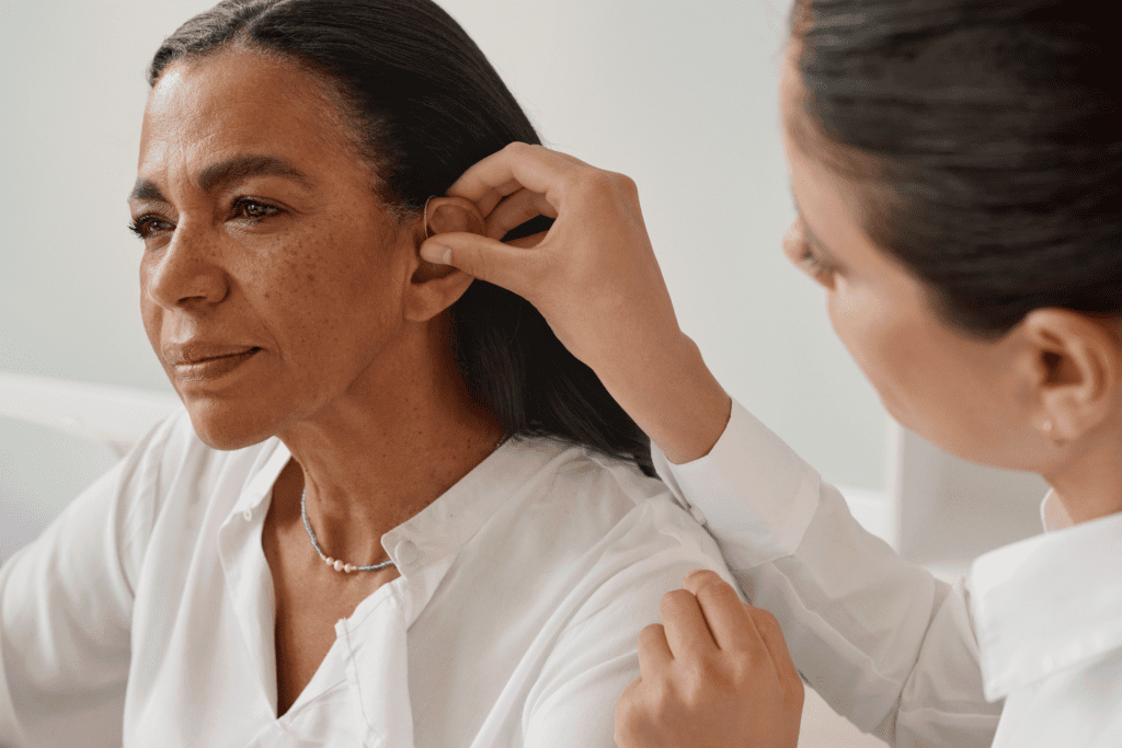 A woman has a hearing aid adjusted by another person, both wearing white shirts, in a bright indoor setting.
