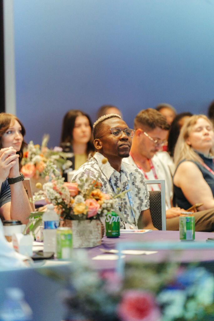 People sit at tables with flowers and drinks, attentively listening during an indoor event or conference.