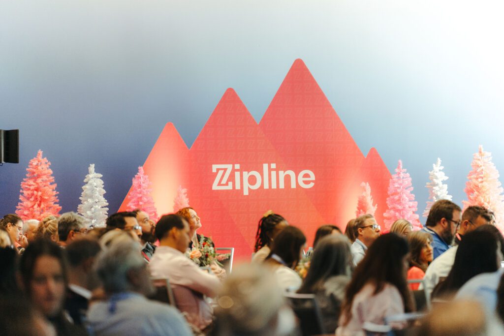 A group of people seated and facing a stage with a red Zipline logo and stylized mountain backdrop.