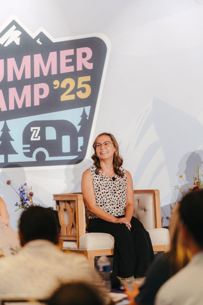 A woman sits and smiles on stage in front of a "SUMMER CAMP '25" sign during an event, with people seated in the audience.