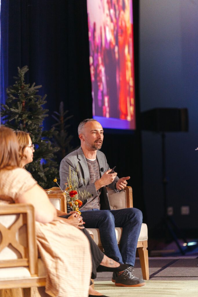 A man speaks while seated on stage next to another person during an indoor event, with a projected image and decorated tree in the background.