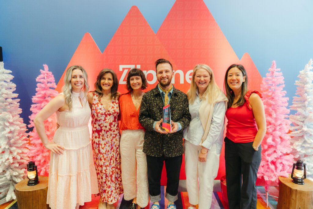 A group of six people stand together smiling, with one person in the center holding an award. They pose in front of a red and blue backdrop with white decorative trees.