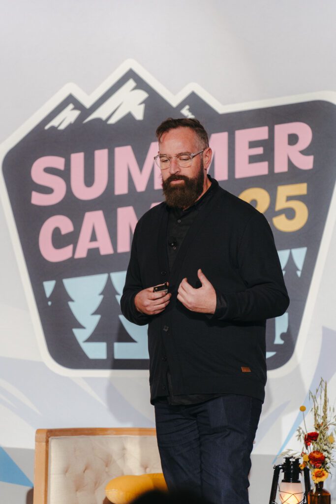 A bearded man wearing glasses gives a presentation onstage in front of a "Summer Camp 2025" sign.