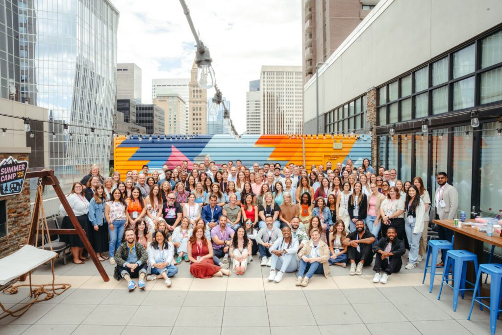 A large group of people pose together on a rooftop patio in an urban setting, with colorful wall art and tall buildings in the background.
