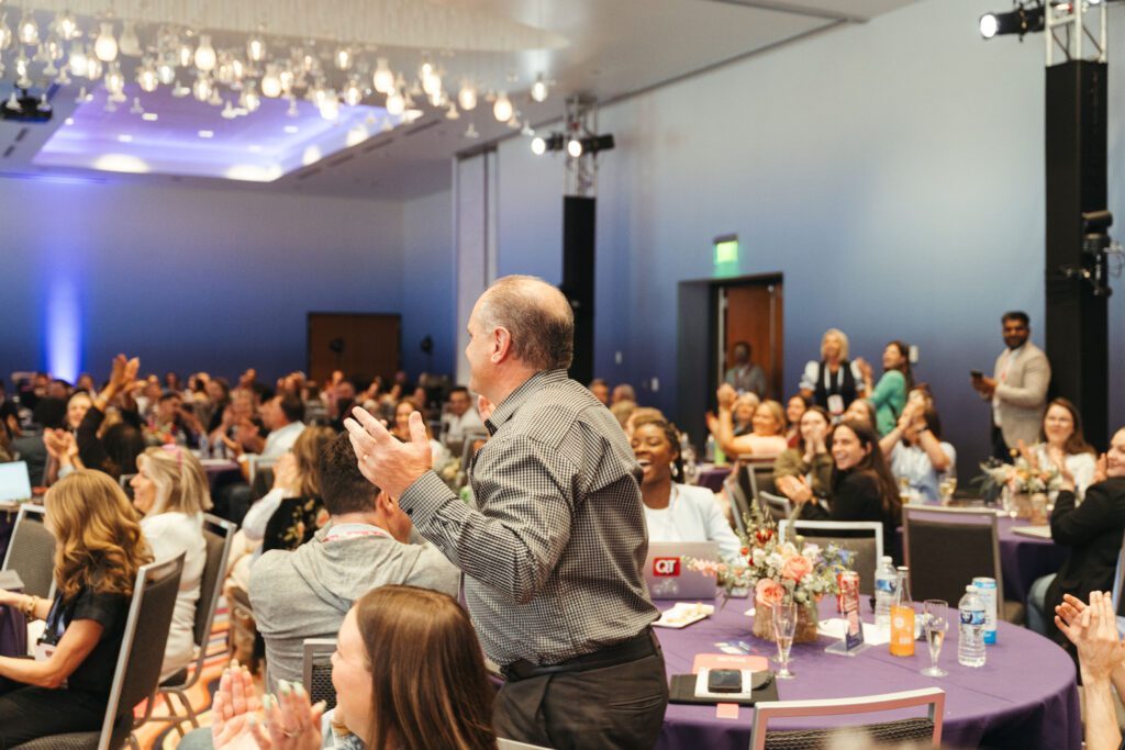 A group of people sit at round tables in a conference room, some standing and applauding while others smile and watch.