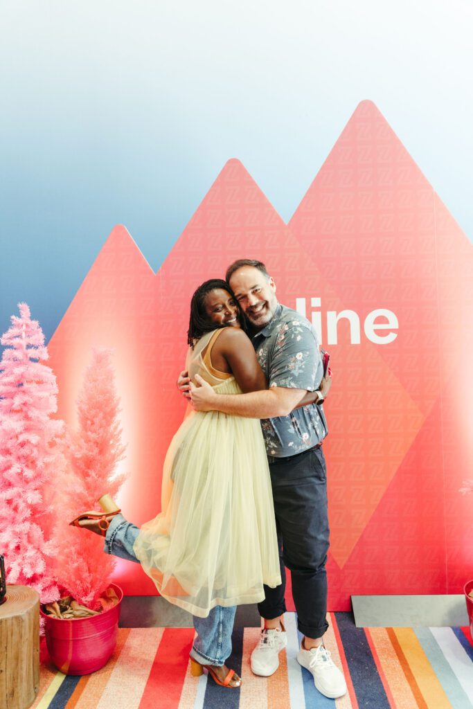 Two people stand smiling and hugging in front of a red geometric backdrop with the partial word “line.” Pink artificial trees and colorful striped carpeting are visible.
