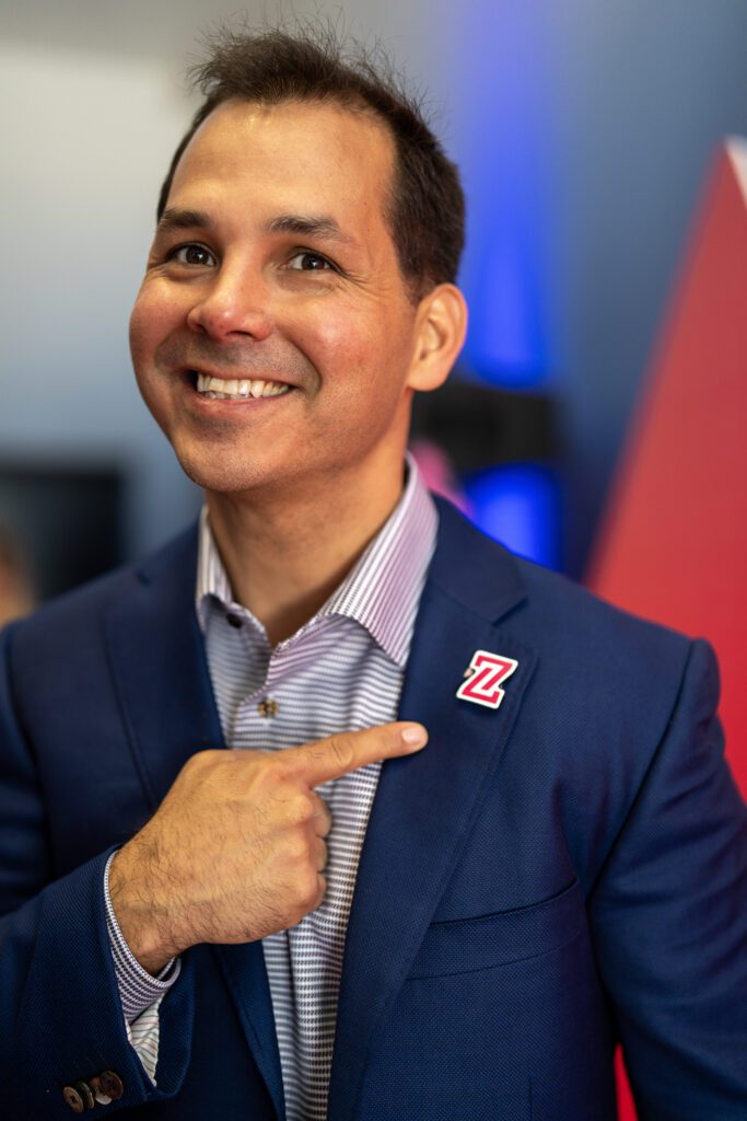 A man in a blue suit smiles and points to a pin with a red "Z" logo on his jacket lapel.
