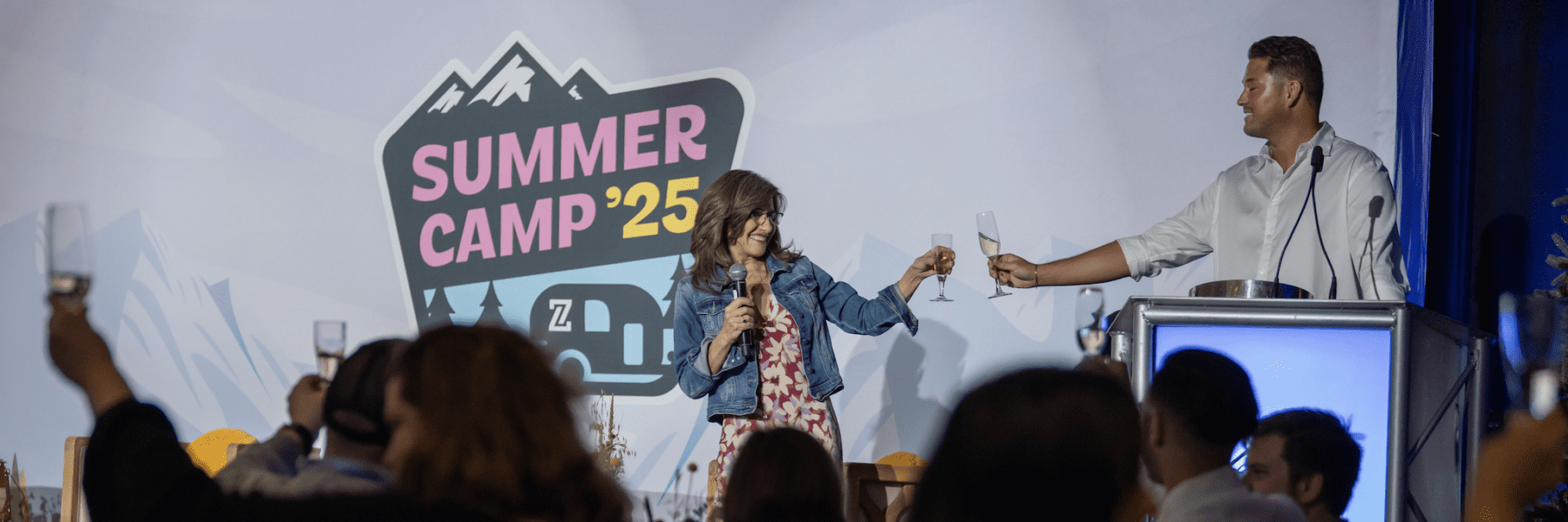 Two people on stage clink glasses at a podium during a "Summer Camp '25" event, while the audience raises glasses in a toast.