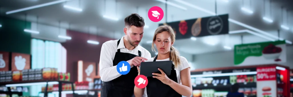 Two supermarket employees in black aprons look at a tablet, with thumbs up, thumbs down, and megaphone icons floating around them in a grocery store aisle.