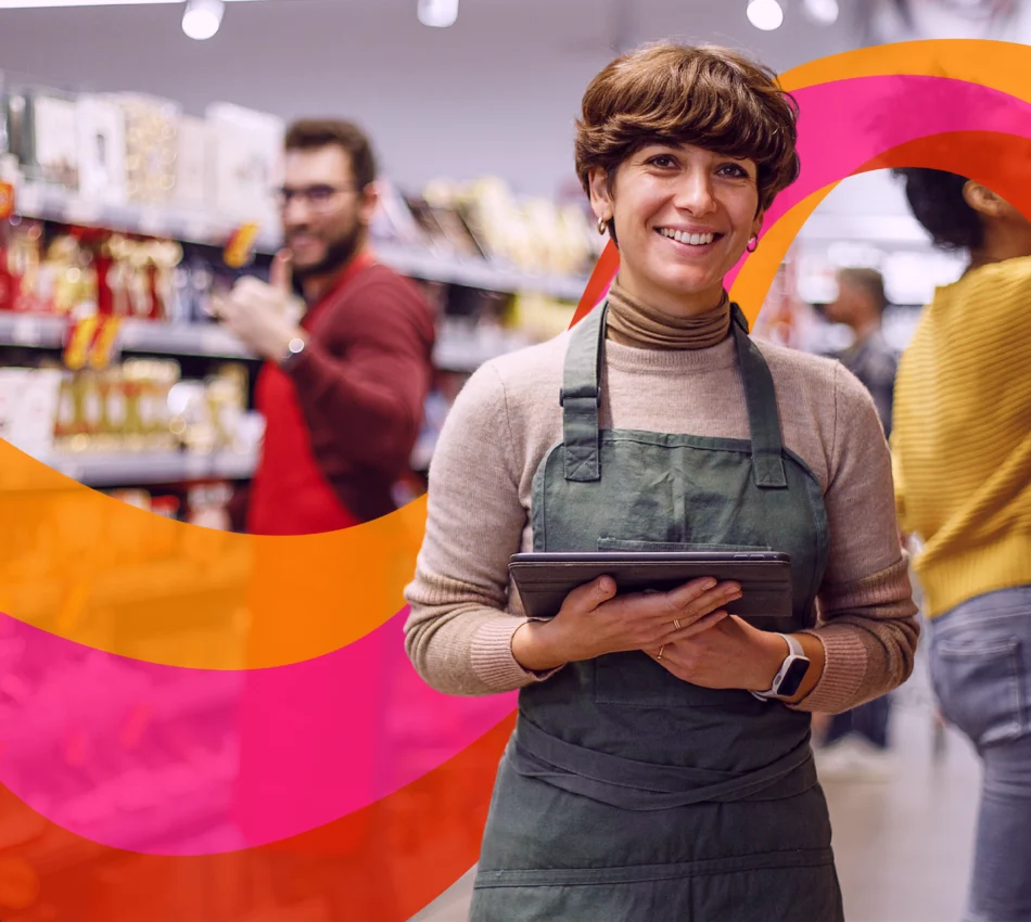 A smiling retail worker in a green apron holds a tablet in a store aisle, with two other people and shelves of products in the background.