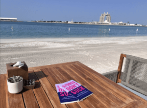 Wooden table with two books, a small potted plant, a napkin box, and a pepper shaker set on a sandy beach, with calm blue water and a large hotel in the background.