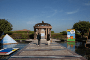 Two people walk down a stone path over water toward a gazebo, surrounded by modern art installations, trees, and clear blue sky.