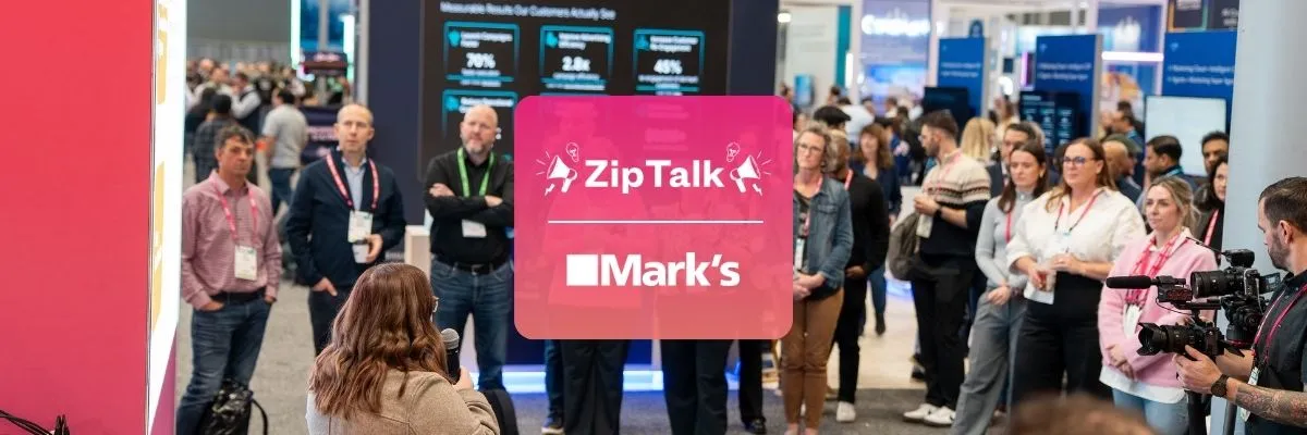 A group of people stands and listens to a speaker at a conference or expo. A large screen and a sign reading "Zip Talk Mark's" are visible in the background.